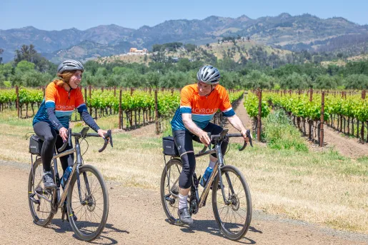 Man and woman wearing orange biking gear, riding bikes in front of a field of crops