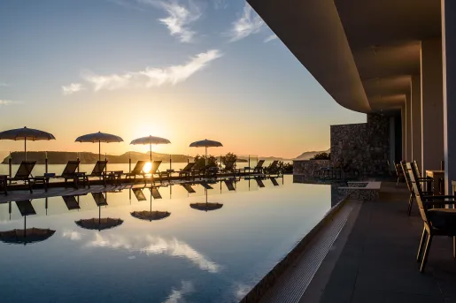 Outdoor pool, surrounded by reclining chairs and umbrellas, with the sunset in the distance