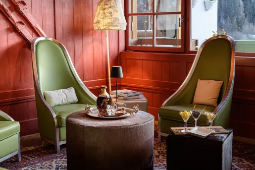 Room with red, wooden walls with green cushioned chairs and an ottoman with a bottle of liquor on top