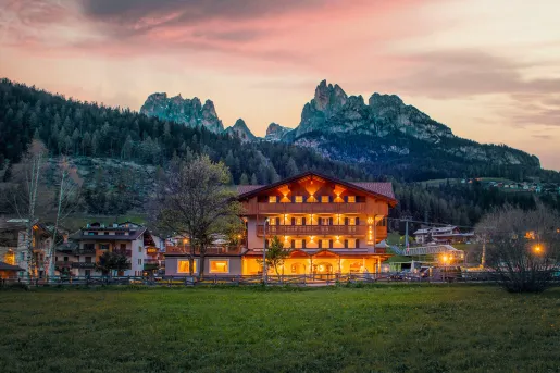 Exterior view of hotel illuminated by warm lights, with a forest and tall mountains in the background