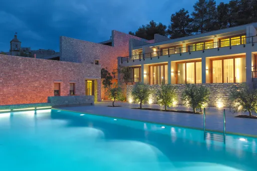 Night time view of an outdoor pool, with an illuminated building with large, glass walls and mini balconies