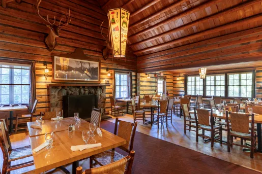 Lodge-style restaurant dining hall with wooden tables and chairs, with a deer head hanging over a fireplace