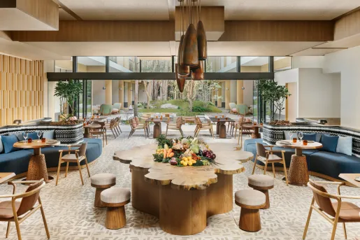 Dining hall with large wooden tables and cushioned chairs, with an outdoor garden in the background