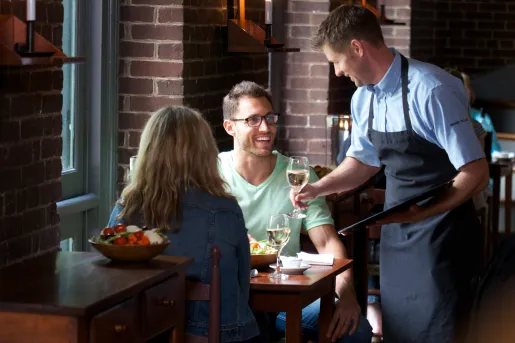 Man and woman sitting at a dining table, with a waiter handing them glasses of wine