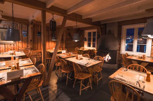 Dimly lit cabin restaurant with dining tables and wooden chairs, with a fireplace in the back
