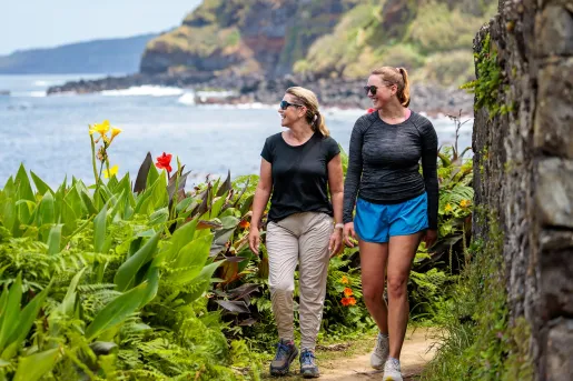 Two women smiling while walking on a dirt trail surrounded by large plants