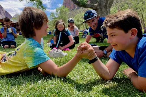 Two kids laying down on the grass, arm-wrestling
