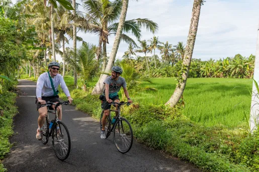 Two men riding bikes on an empty road with a large grass field and palm trees