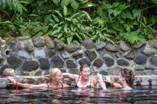 Three women holding drinks and swimming in a pool, surrounded by stone walls