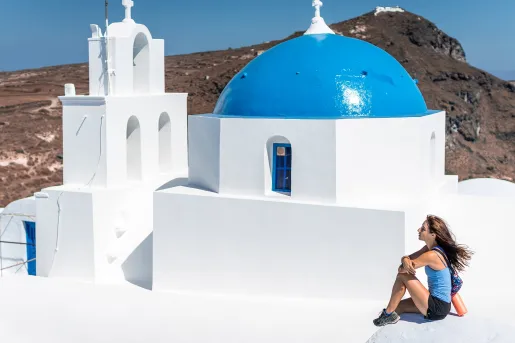 Woman sitting on the roof of a white, Greek house, with large cliffs in the background