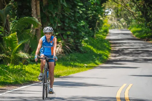 Woman wearing a blue and orange jersey, riding a bike on a road surrounded by tall plants