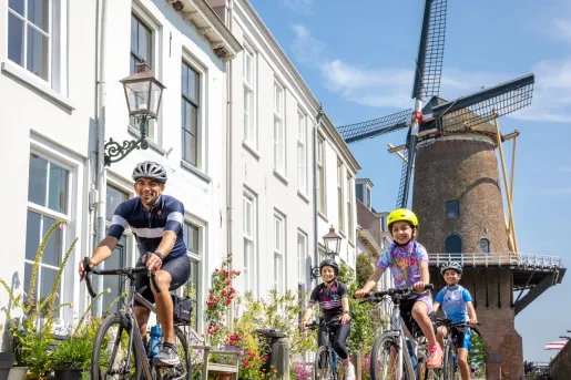 Man and group of kids riding bikes on a road, with a large windmill in the background