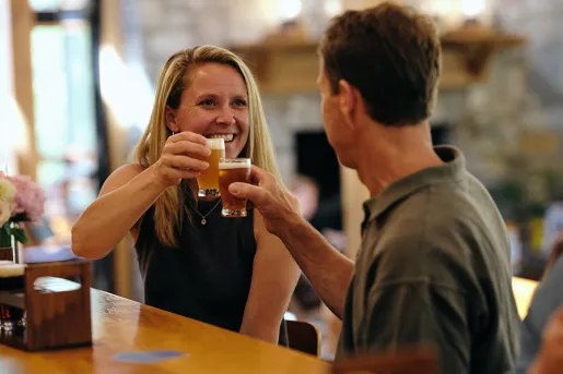 Man and woman sitting at a bar, smiling while raising glasses of beer