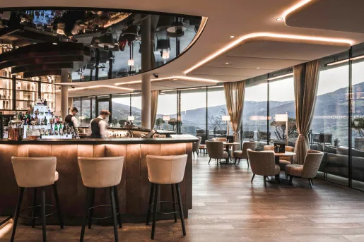 Indoor bar and lounge with two bartenders behind the counter, with beige stools and wooden tables in the room