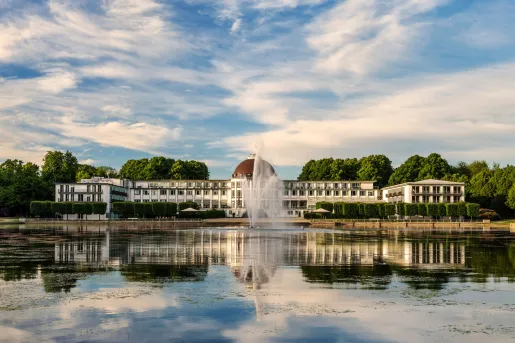 Wide, exterior view of white building with brown dome in the center, with a large fountain in front