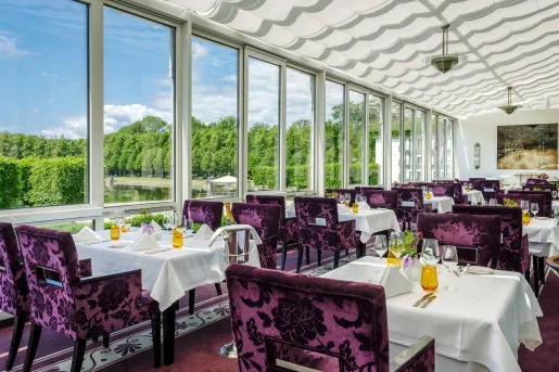Restaurant dining area with purple velvet chairs and white tables, with large windows along the walls