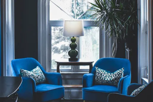 Room with two blue, cushioned chairs next to a small wooden table with a lamp on top