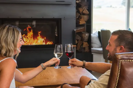Man and woman smiling in front of a fireplace, holding up glasses of wine