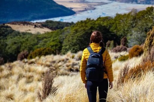 Woman in a yellow jacket walking on a gravel trail, surrounded by tall weeds