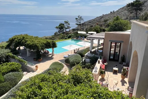 Outdoor pool and beige hotel building, surrounded by trees and views of the ocean in the distance