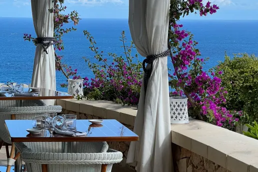 Outdoor dining area with wooden tables and woven white chairs, with the ocean in the background