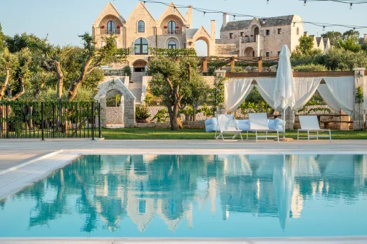 Outdoor pool, surrounded by white chairs and umbrellas, with large stone buildings in the distance