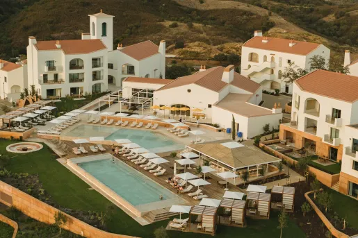 Sky view of white hotel buildings with outdoor pools and tall hills in the background