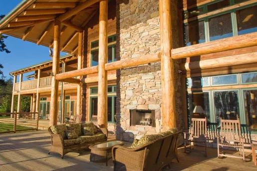 Cabin building exterior with large wooden pillars and a stone fireplace, with cushioned chairs in front