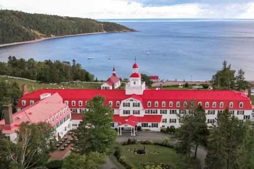 Exterior view of long white building with a red roof, with a large garden in front and the ocean in the distance
