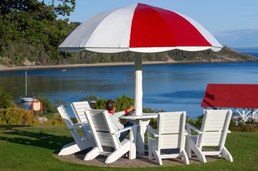 Person sitting on a white lawn chair in front of a red and white table, looking out to the ocean