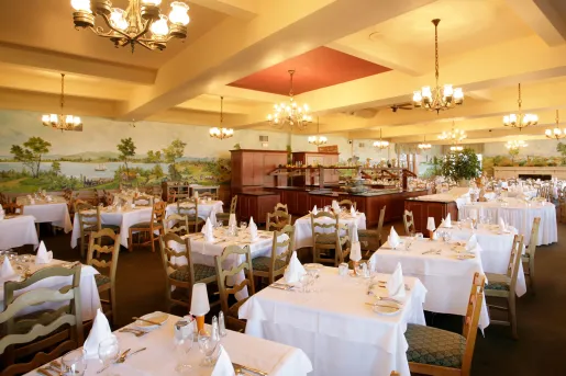Restaurant dining hall with cushioned chairs and white cloths over dining tables, with a large mural along the wall