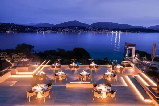 Illuminated outdoor patio with woven chairs and white, circular tables, looking out towards a large lake
