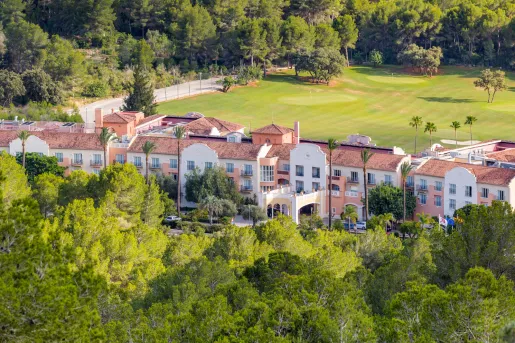 Long, beige and brown hotel building with a large grass field in the back and tall trees in front