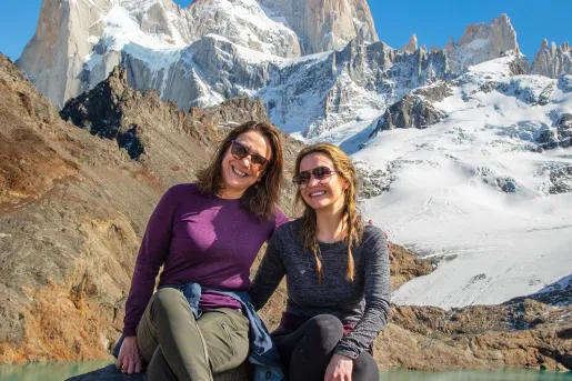 Two women smiling while sitting on boulders in front of a lake