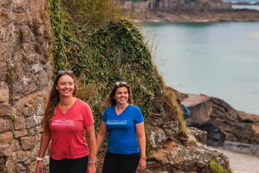 Two women walking along a stone back with hills and the ocean in the background
