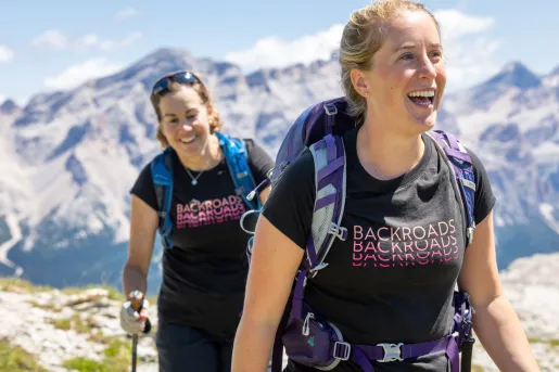 Two women smiling, wearing backpacks while hiking on a mountain