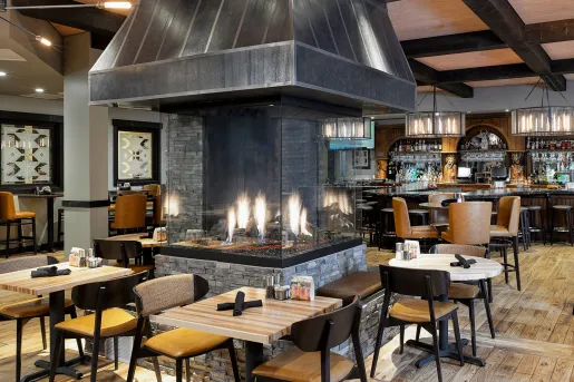 Restaurant dining area with wooden chairs and tables, with a large, enclosed firepit in the center