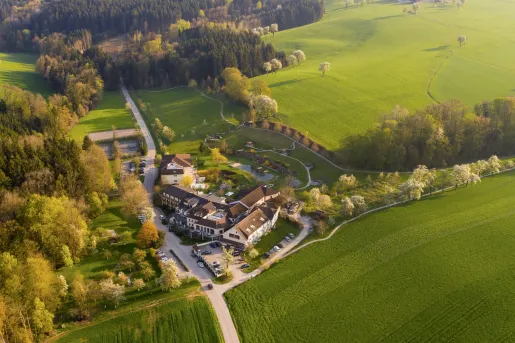 Sky view of large hotel building in the middle of a grassy field, surrounded by a forest