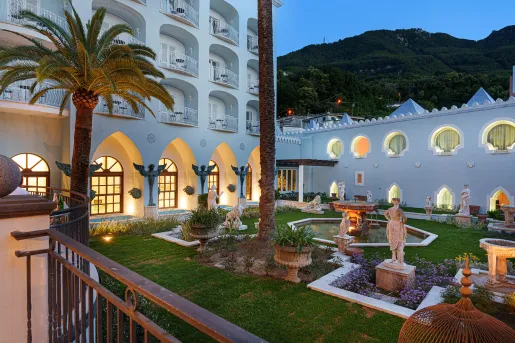 The interior garden of Palazzo Manzi Terme & Spa in Casamicciola Terme on the island of Ischia, Italy features manicured gardens, classical white marble statues, a stone fountain, and large palm trees.