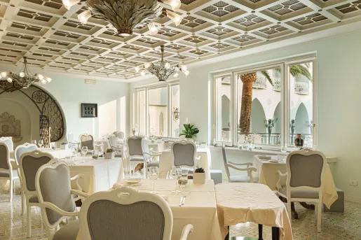 The dining room at the Palazzo Manzi Terme & Spa in Casamicciola Terme on the island of Ischia, Italy features white armchairs, linen-draped dining tables, and a distinctive coffered ceiling accented with decorative chandeliers.