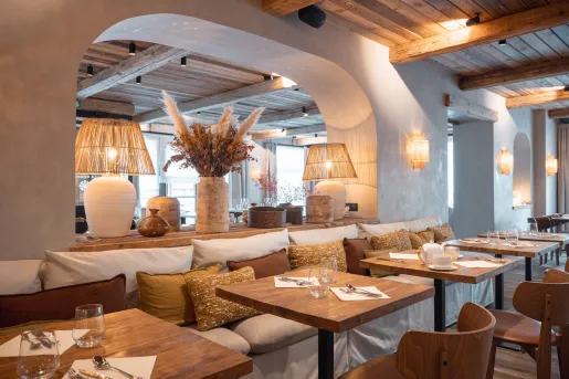 Elegant hotel restaurant with wood-beam ceilings, natural wood furnishings, woven pendant lamps, and cushioned banquette seating. Decorative vases and warm lighting create a refined yet cozy dining atmosphere.