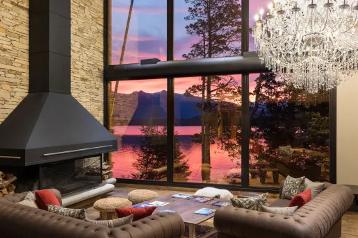 Calfuco Wine Hotel & Spa features a grand, double-height main living lounge framing the shifting sunset colors of Patagonia. The room incorporates woven rattan floor poufs, a solid slab wooden coffee table, plush couches, and an exposed wood pile for the hearth.