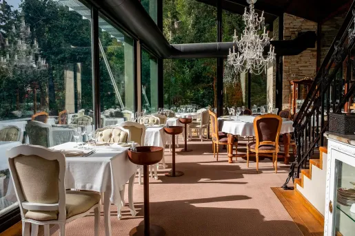 Fine-dining restaurant area inside Calfuco Wine Hotel & Spa featuring white linens, elegant chandeliers, and floor-to-ceiling glass windows that look out directly into the bright green Patagonian forest in Neuquén, Argentina.