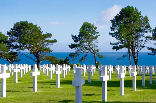 Rows of white cross-shaped headstones stretch across a manicured lawn at a coastal military cemetery, with pine trees and the blue sea visible in the background.