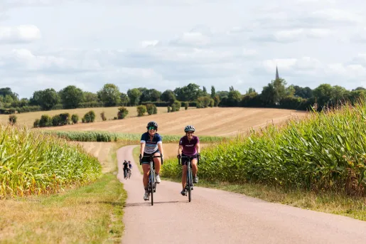 Two cyclists ride along a quiet country road bordered by tall cornfields, with rolling farmland, hedgerows, and a distant church spire stretching across the rural landscape.