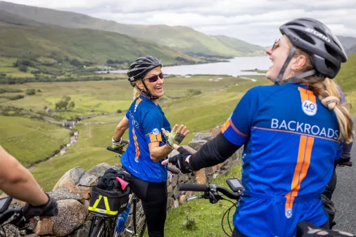 Two women smiling while walking with their bicycles, looking out towards a large valley