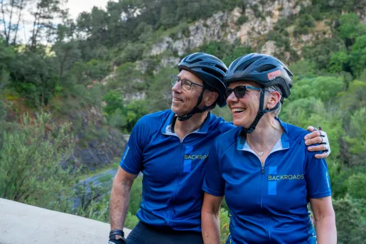 A happy couple wearing blue cycling jerseys with the "BACKROADS" logo, helmets, and sunglasses pause on a scenic overlook.