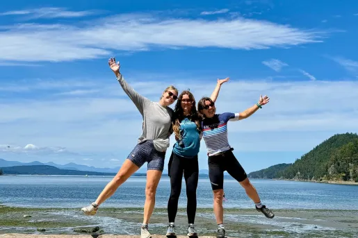 Three guests smiling with hands raised while standing on a driftwood log along the a trail in British Columbia.