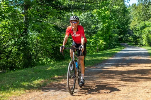 Man smiling while riding a bike on a road covered with leaves, surrounded by tall trees