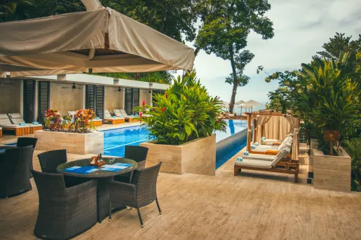 Luxury resort pool area surrounded by tropical landscaping, lounge chairs, and private cabanas. Outdoor dining seating sits beside the pool, with views toward the ocean through the trees in the background.